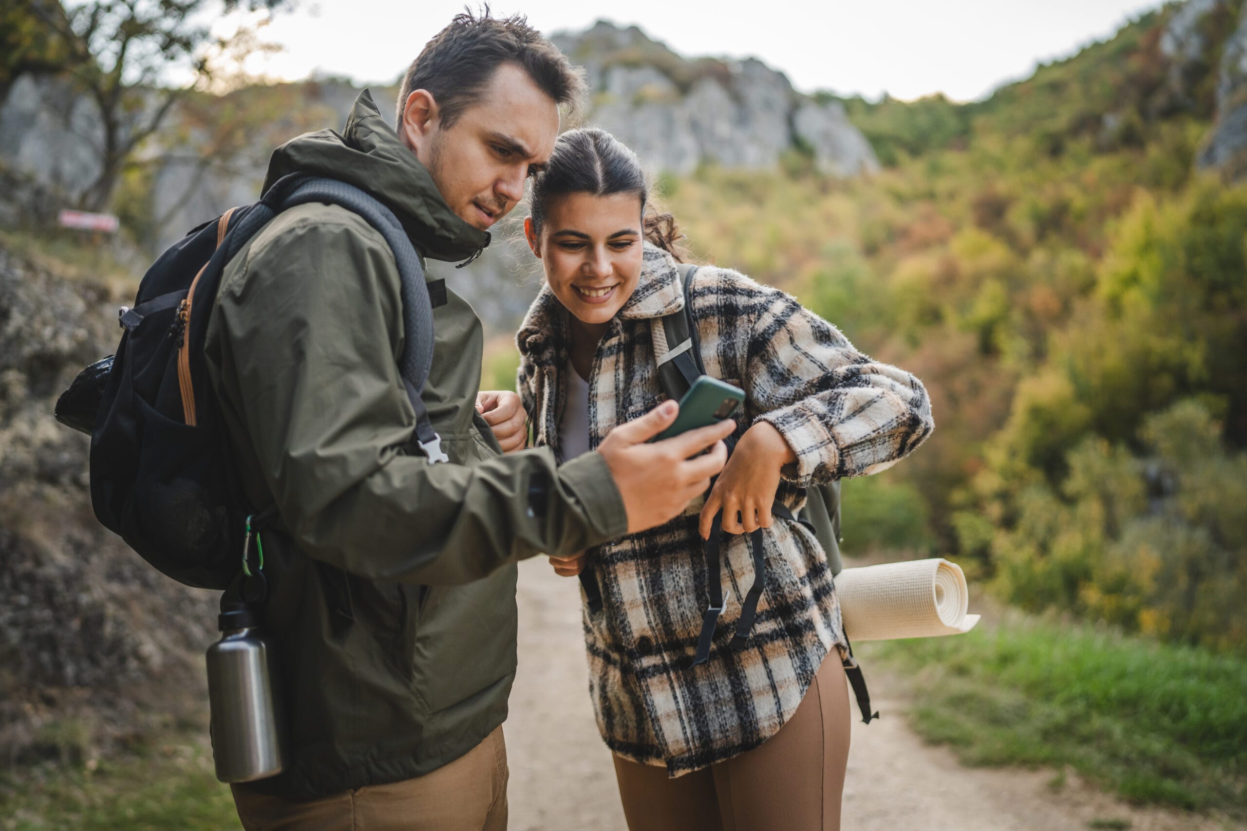 Two hikers share a moment, looking at a smartphone, surrounded by a scenic, colorful landscape.