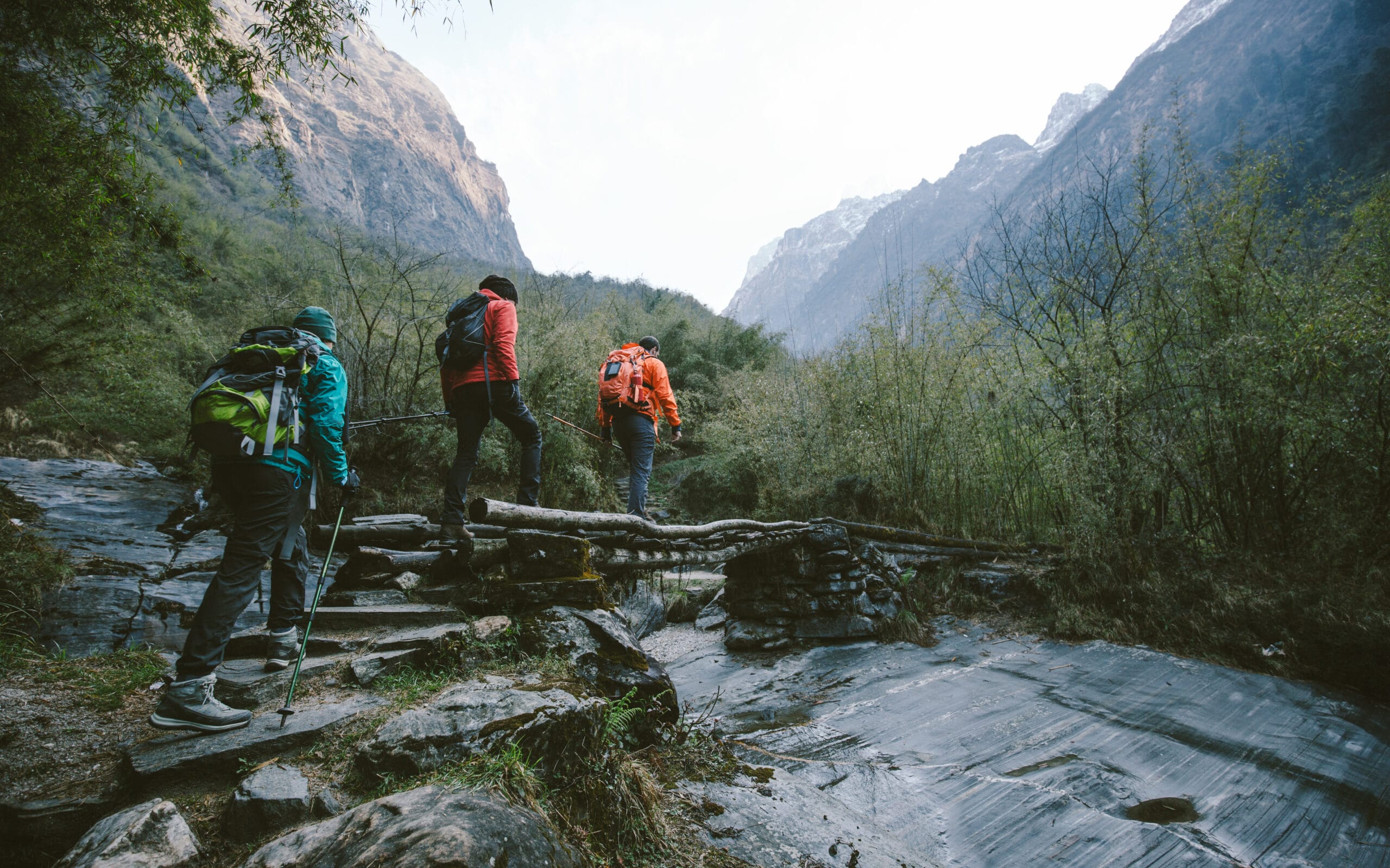 Hikers crossing a wooden bridge in a mountainous landscape, surrounded by lush greenery and steep cliffs.