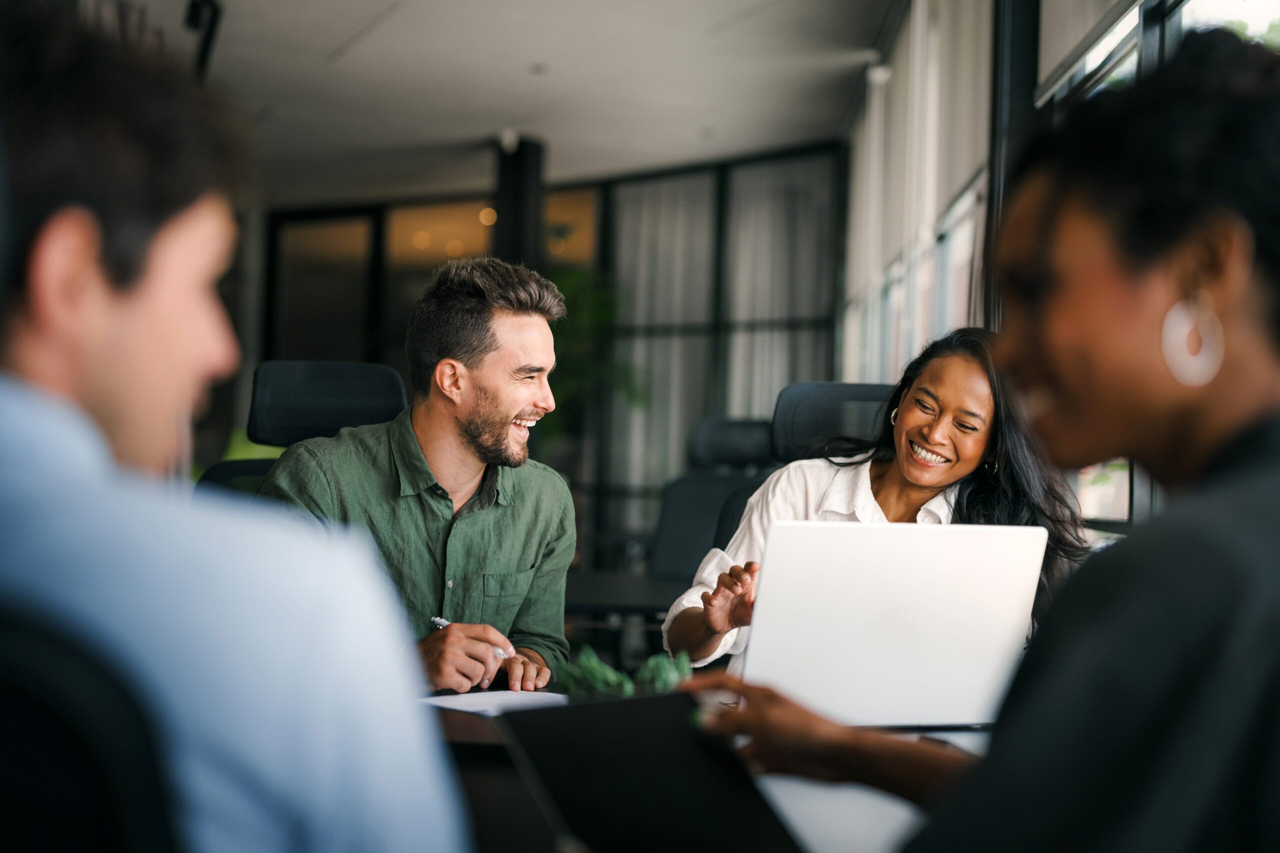 A group of four people in a bright office, engaged in a lively discussion, smiling and sharing ideas over a laptop.