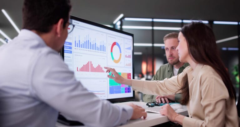 Three professionals analyze data on a large screen, discussing graphs and charts in a modern office setting.