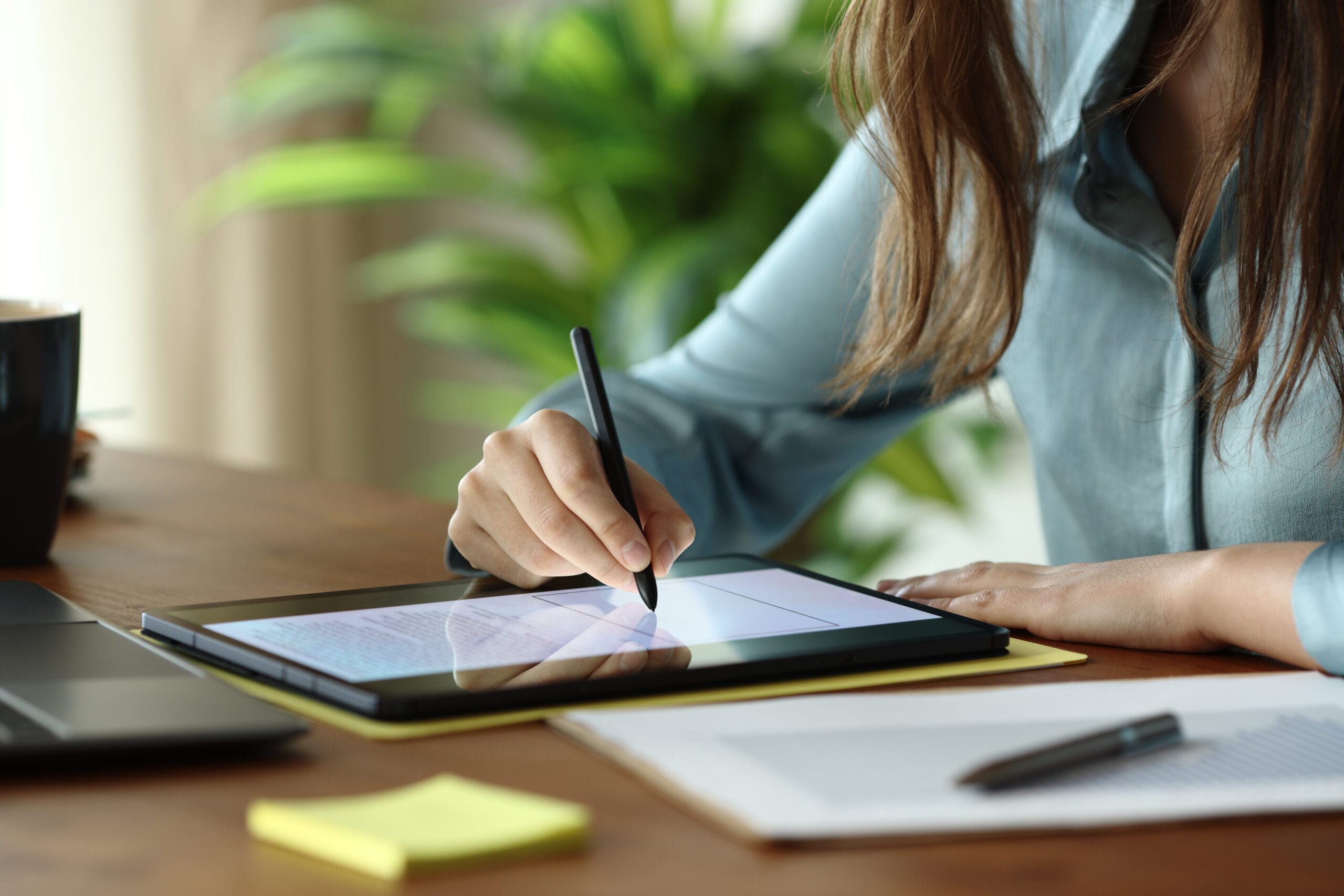A person using a stylus on a tablet, surrounded by a coffee cup, sticky notes, and a laptop, with greenery in the background.