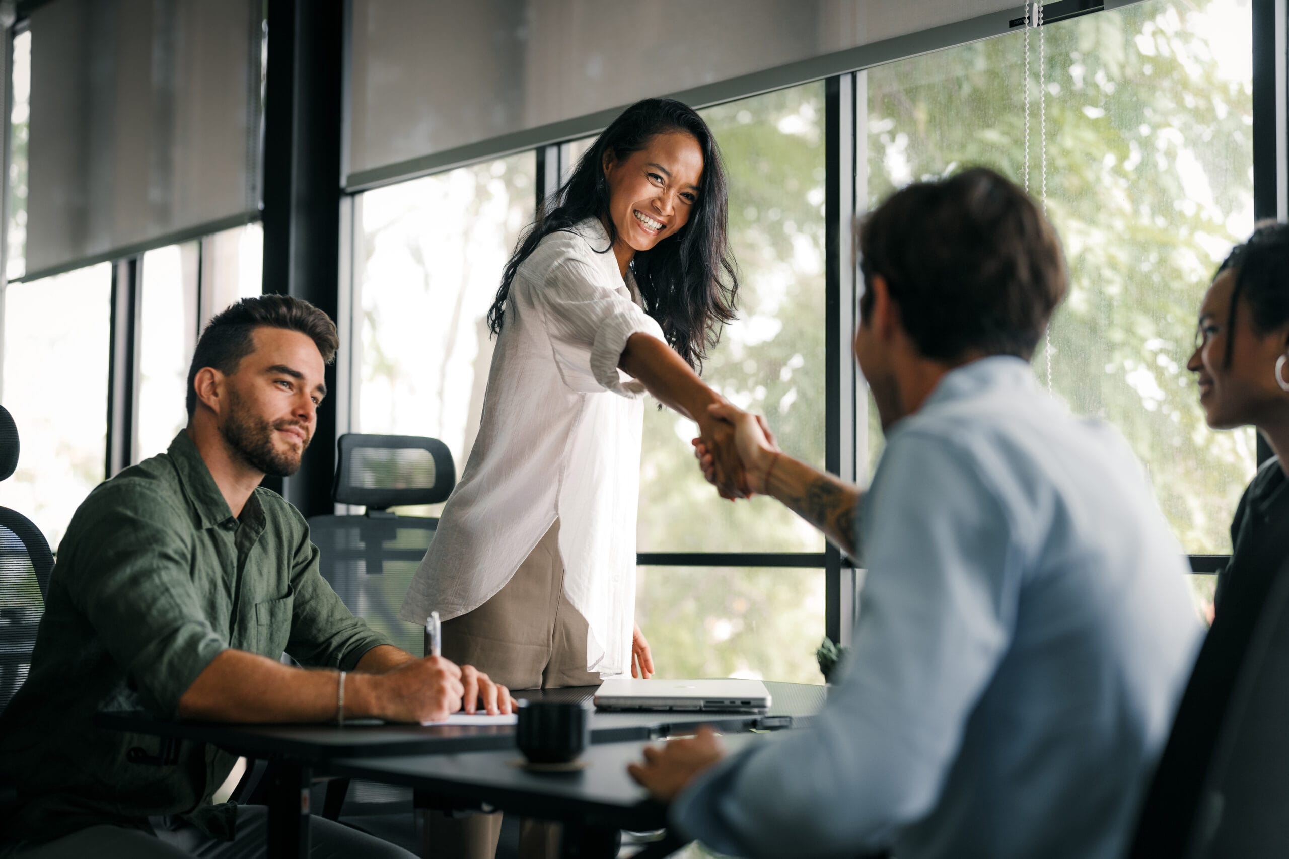 A woman smiles and shakes hands with a man in a modern office, while two others observe at the table.
