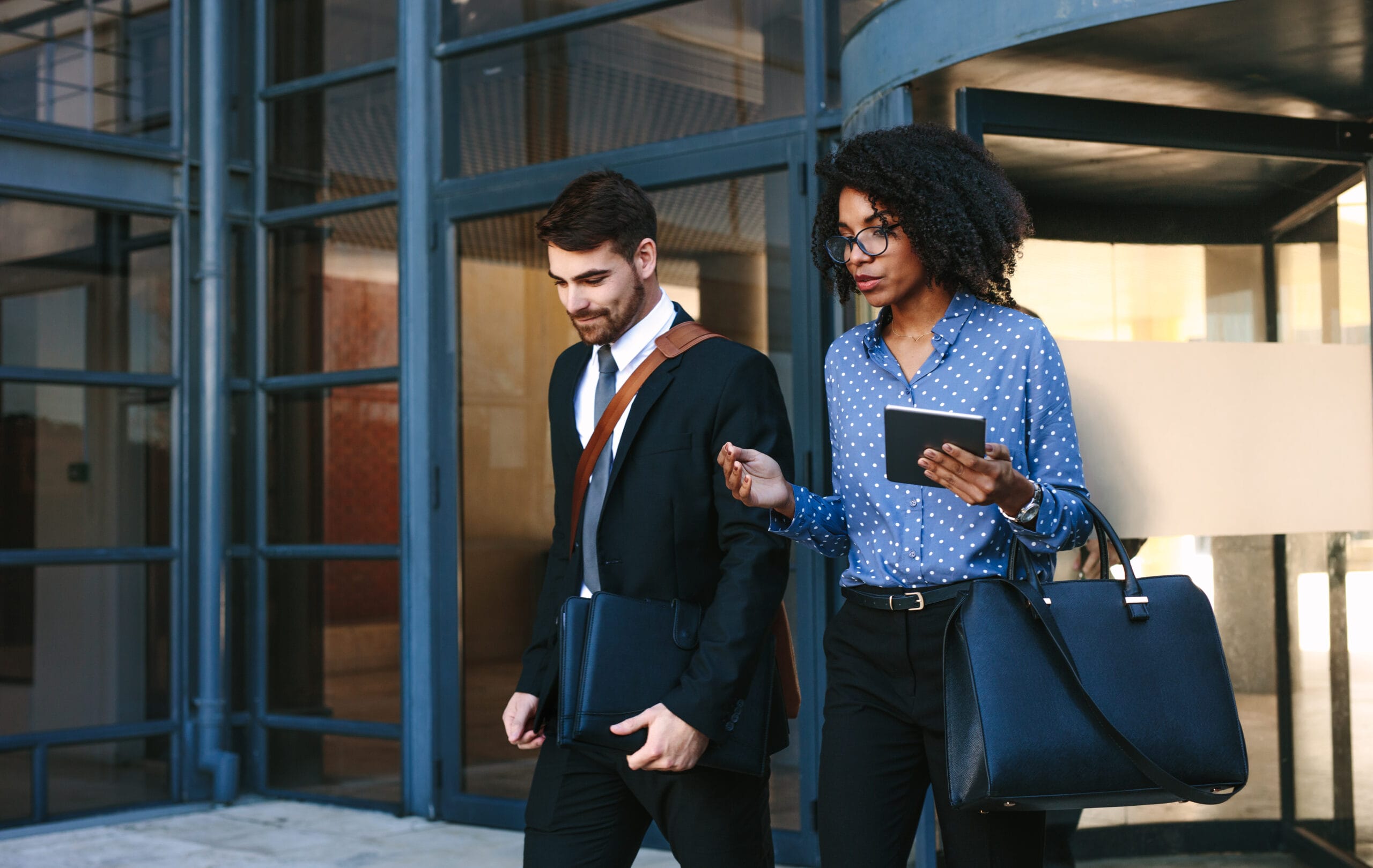 A man in a suit and a woman in a polka dot blouse walk together, discussing while she holds a tablet and a handbag.
