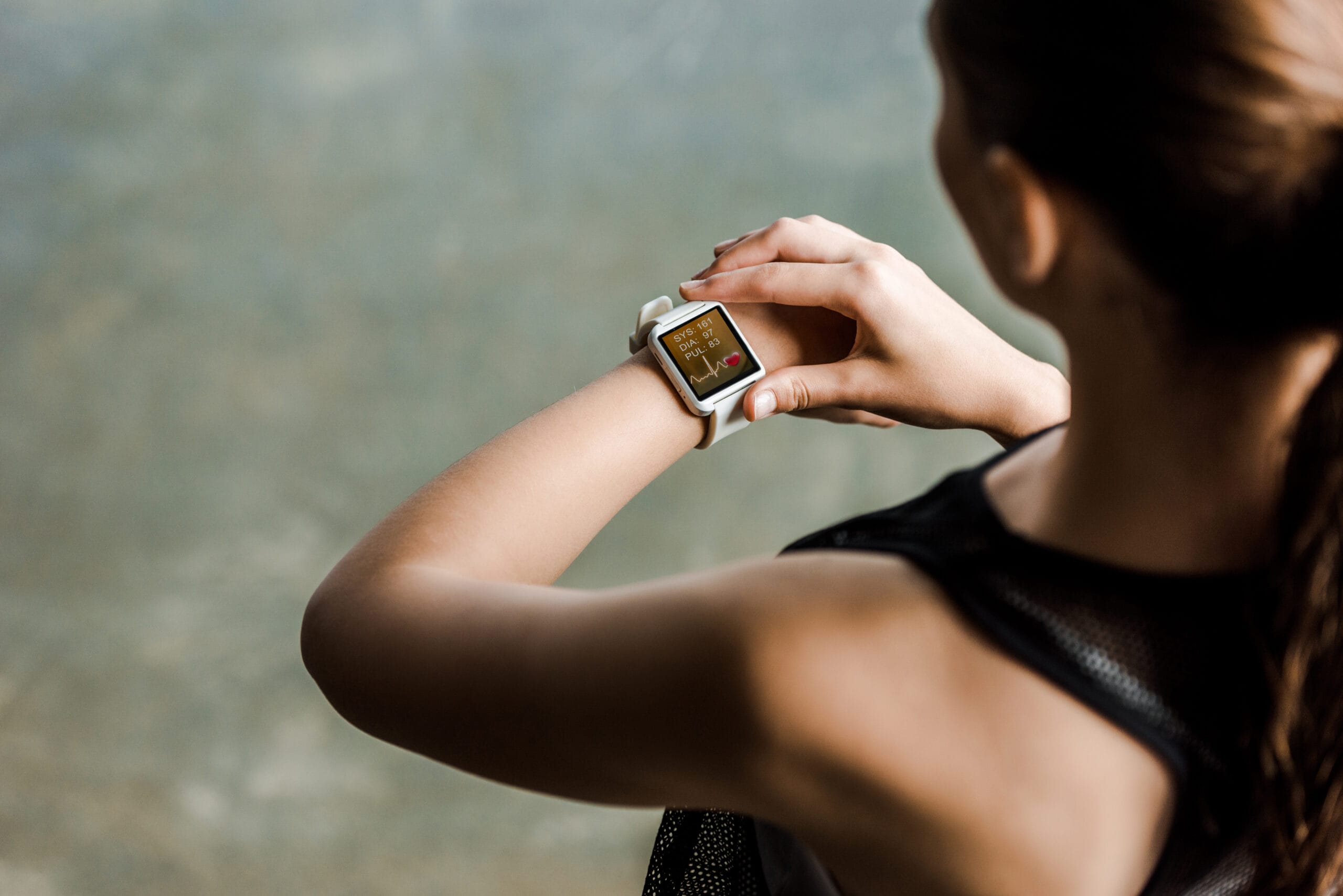 A woman checks her smartwatch displaying health metrics, wearing a sleeveless athletic top against a blurred background.