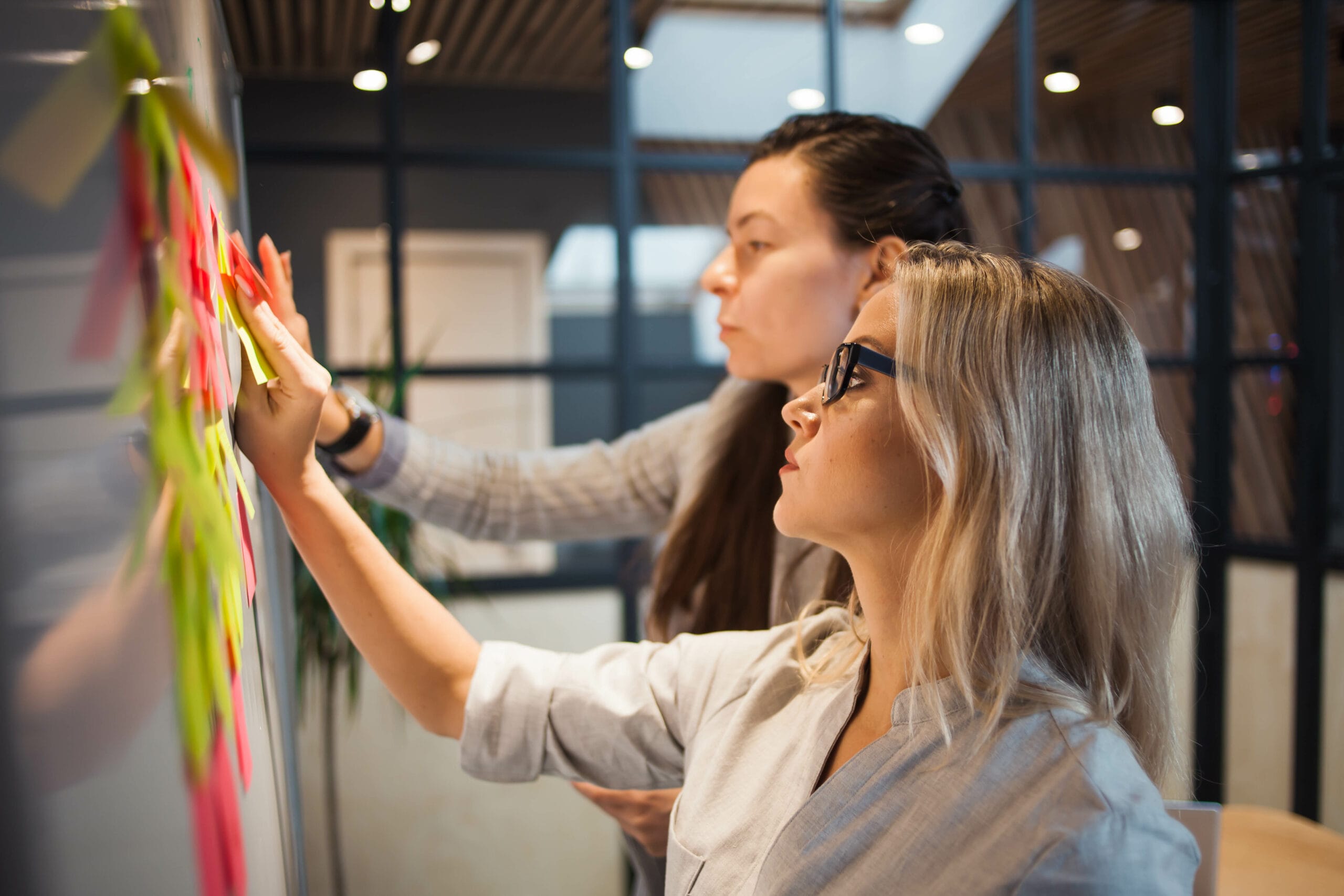 Two women collaborate at a whiteboard, attaching colorful sticky notes in a modern office setting.