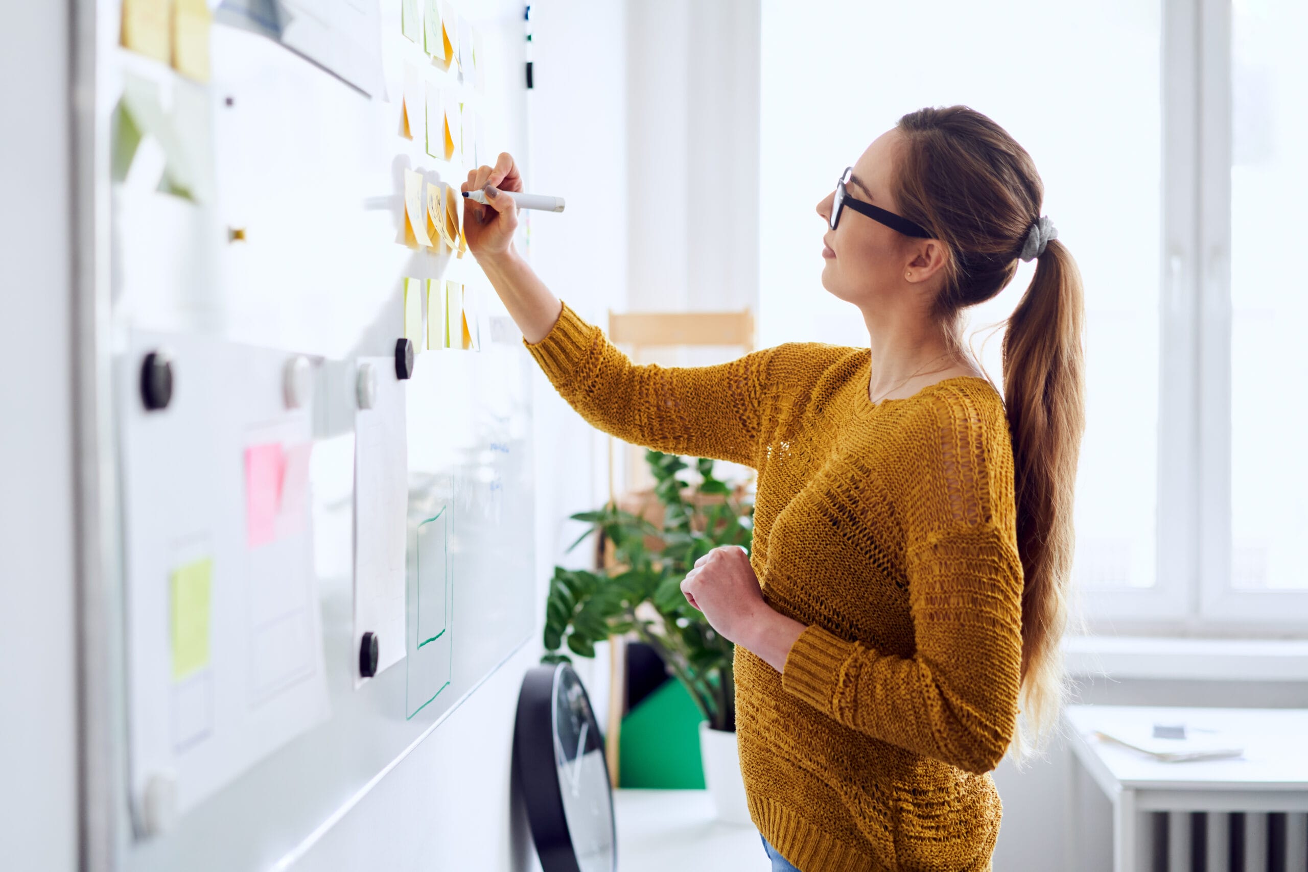 A woman in a mustard sweater writes on a whiteboard filled with colorful sticky notes, in a bright office space.