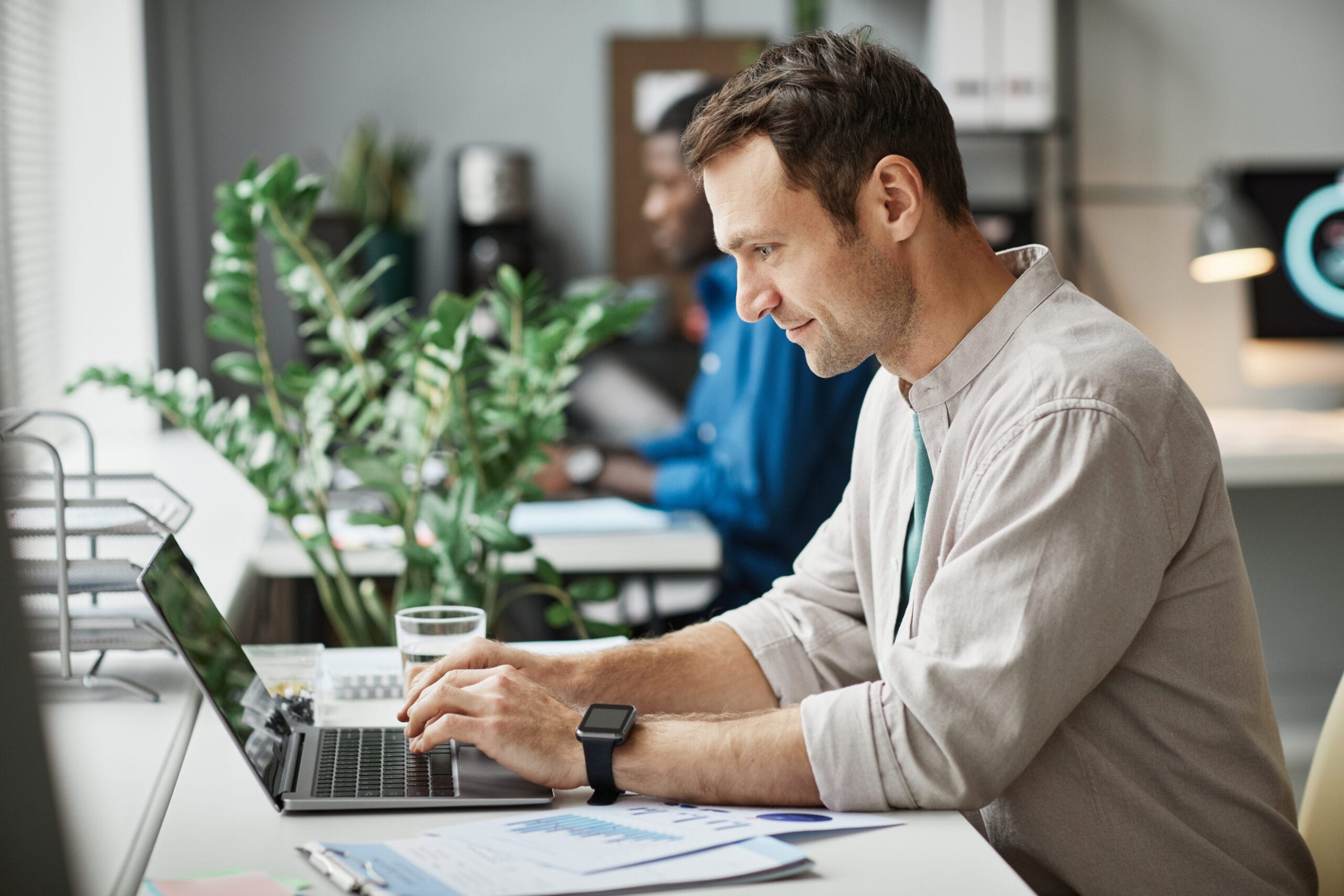 A man types on a laptop at a desk with plants, a glass of water, and documents, while another person works in the background.