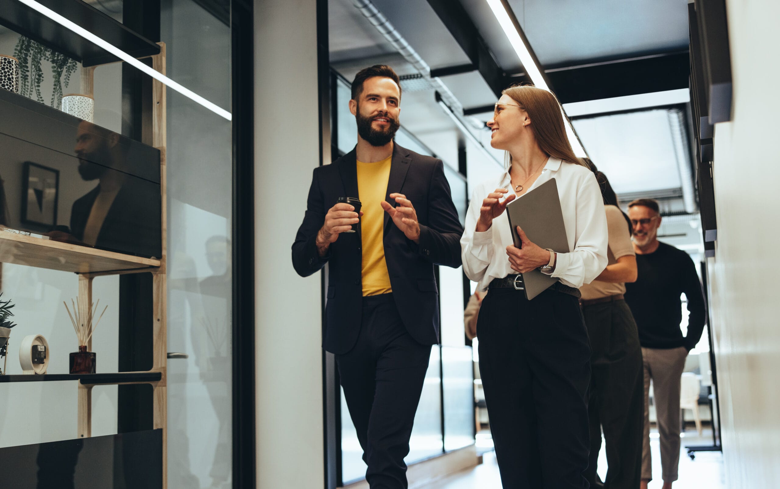 Two professionals engage in conversation while walking through a modern office corridor, with colleagues in the background.