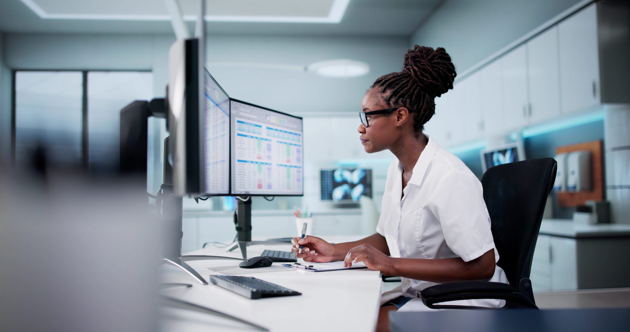 A focused healthcare professional in a white coat analyzes data on dual monitors in a modern medical office.