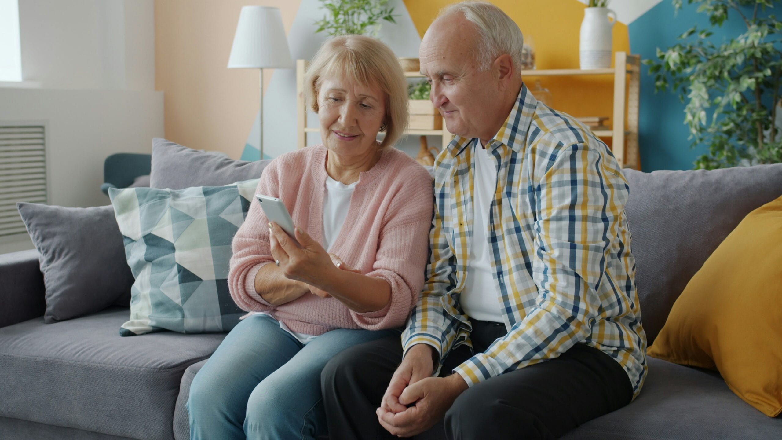An elderly couple smiles while looking at a smartphone together on a cozy couch, surrounded by colorful decor.